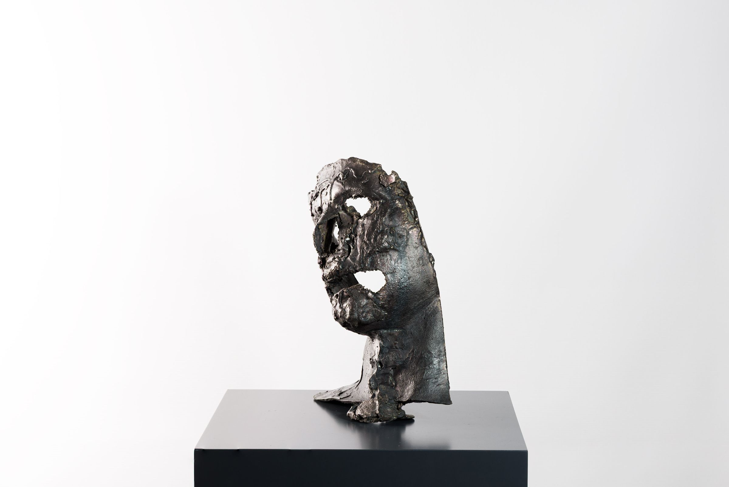 Skulpturel bronzemaske med huller og rå overflade placeret på en sokkel.