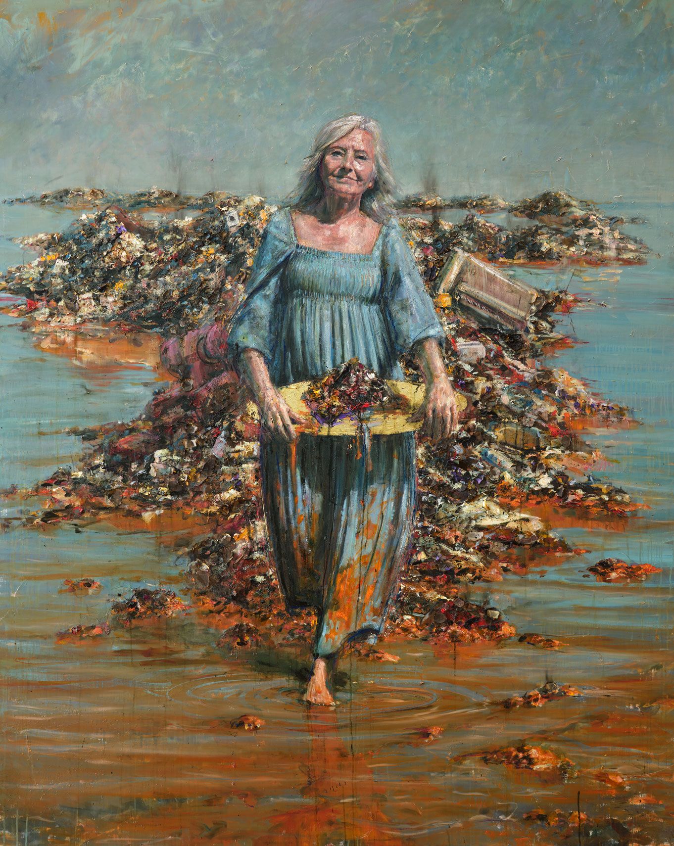 Kvinde går gennem lavt vand foran bunker af affald og bærer en bakke med en mørk masse.