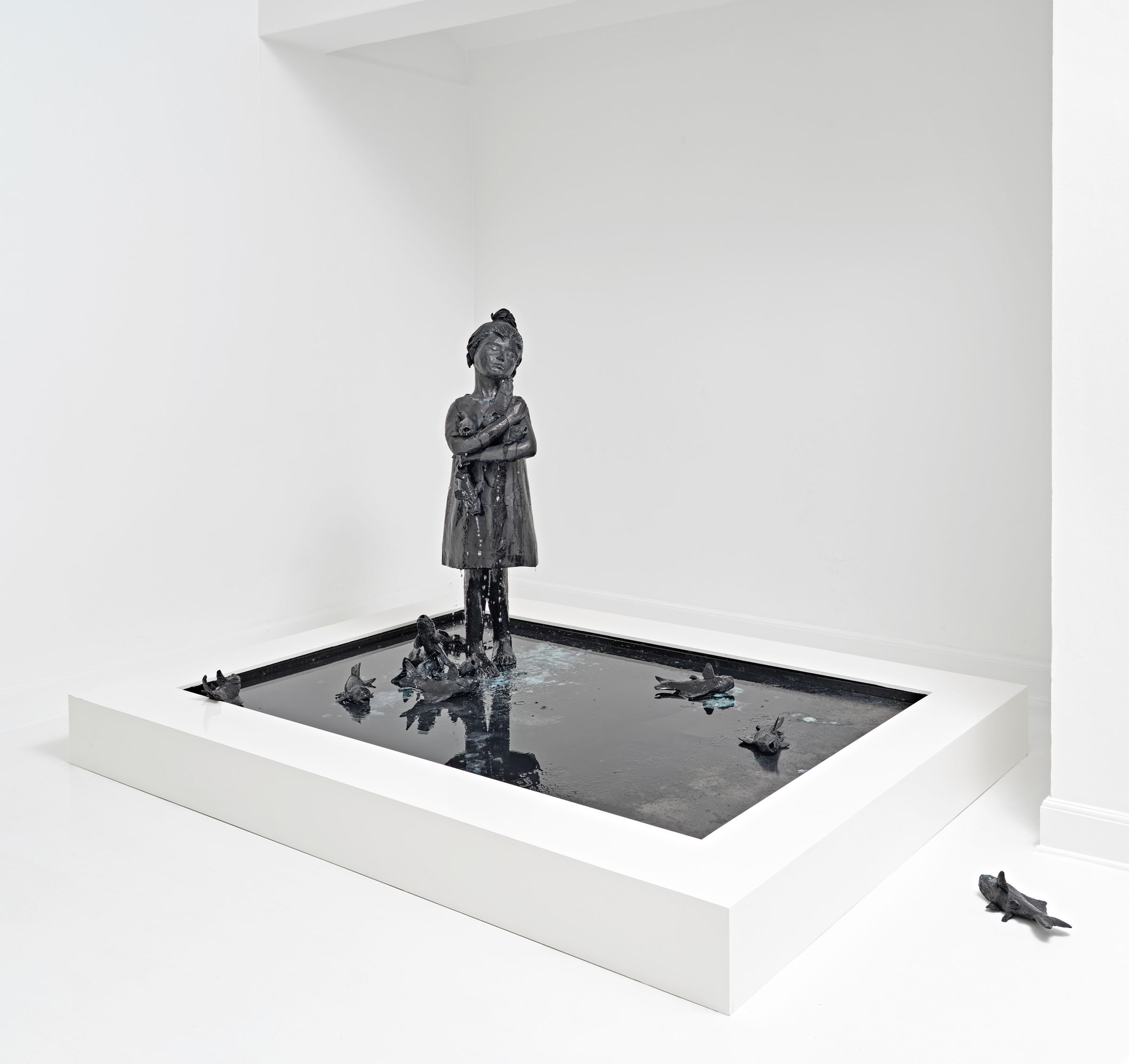 Large bronze sculpture of a girl surrounded by fish in a dark reflective pool.